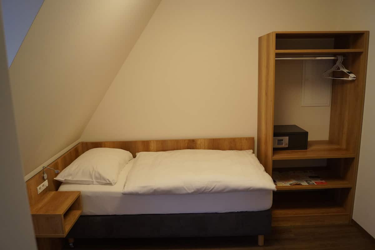 A small, neatly arranged bedroom with a single bed, a wooden nightstand, a built-in wardrobe with a hanging closet, and a shelf with books.
