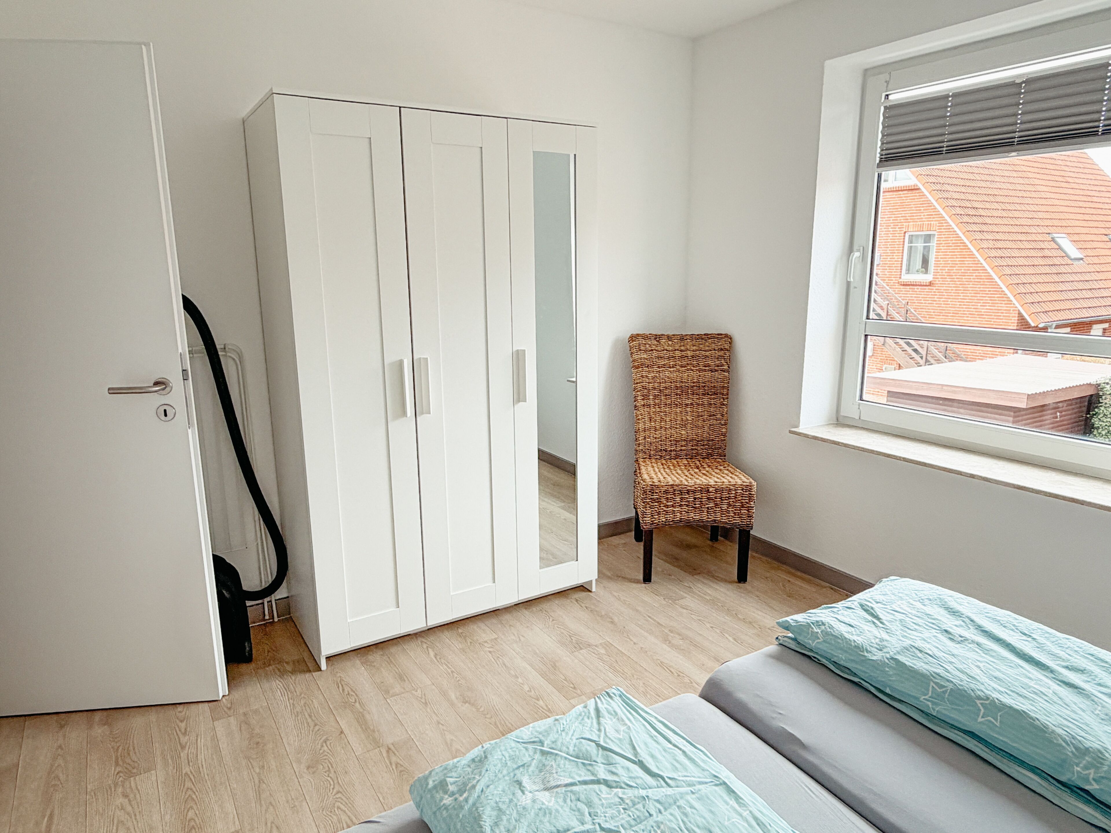 A bedroom with a wardrobe, a chair, and a window with a view of buildings.
