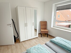 A bedroom with a wardrobe, a chair, and a window with a view of buildings.