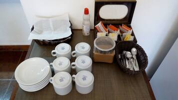 A table with stacked white plates, several white cups with lids, a basket of utensils, a bottle of hand sanitizer, and a box of tissues.