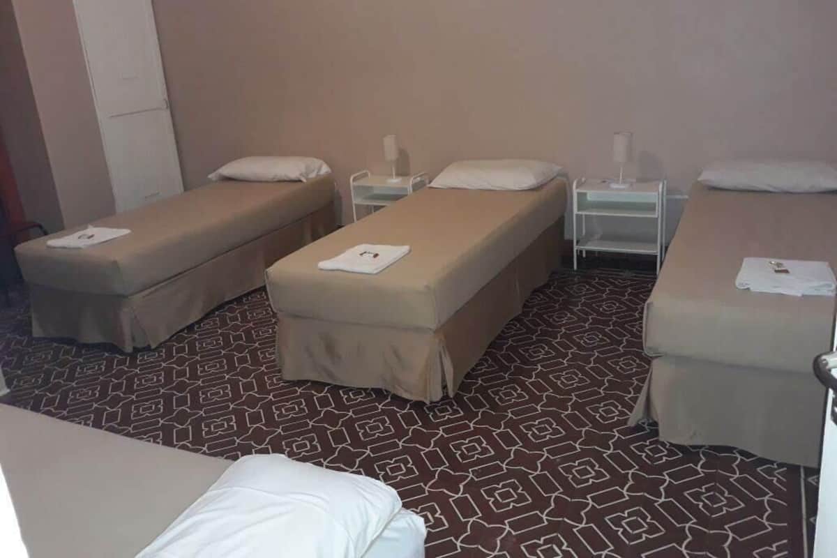 Une chambre d’hôtel avec trois lits simples, chacun équipé d’un oreiller et d’une petite table de chevet. La chambre est dotée d’un tapis à motifs et de murs aux couleurs neutres.