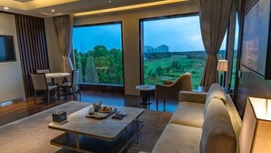 Una sala de estar moderna con un ventanal que ofrece vistas a un paisaje verde y una estructura distante. Hay un sofá, una mesa de centro y un comedor con sillas.
