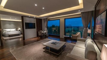 A modern hotel room with a large bed, a seating area with a coffee table, a TV, and a balcony with a city view.