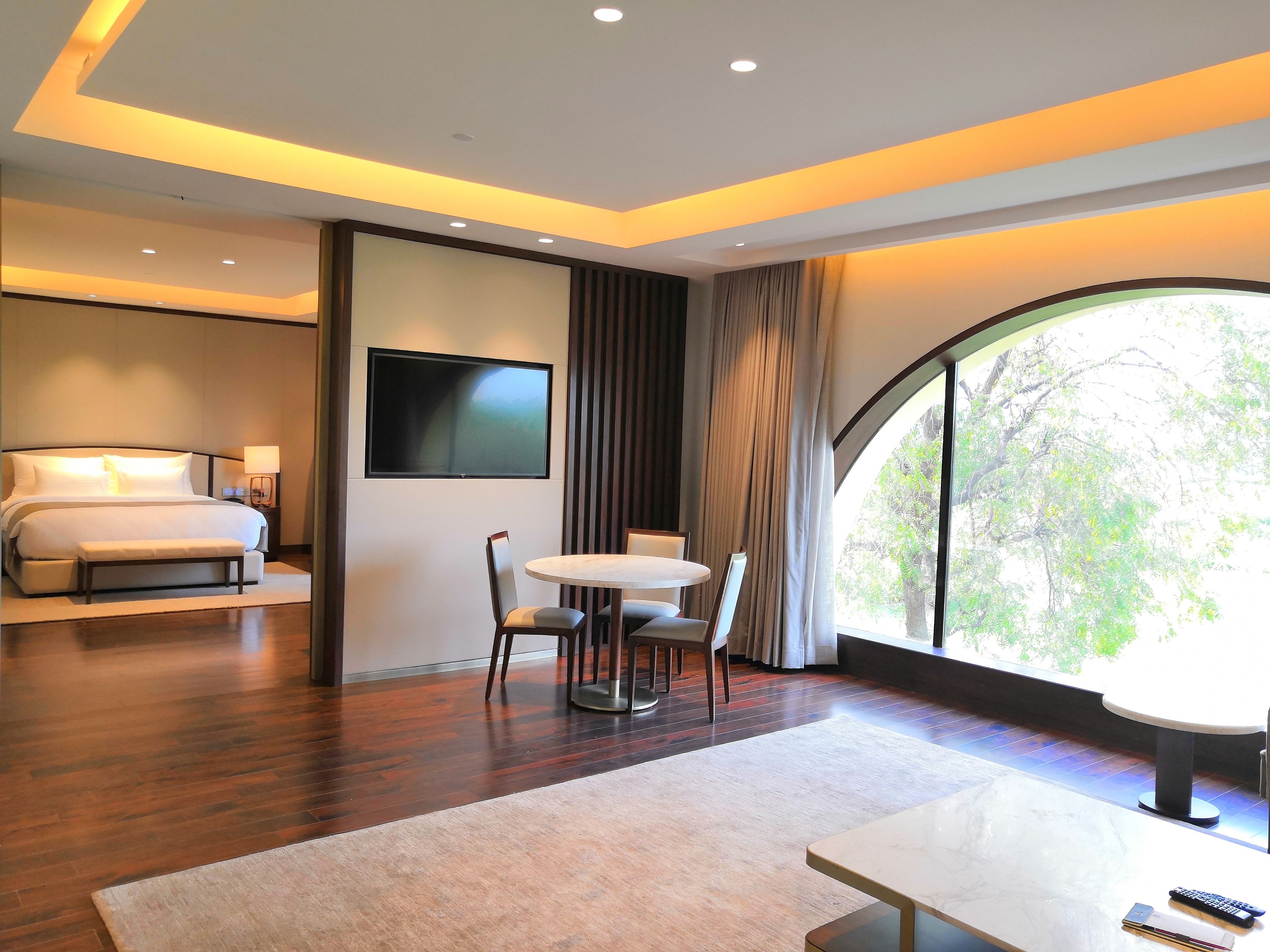 A modern hotel room with a bed, a TV mounted on the wall, a dining table with chairs, and a large window offering a view of trees.