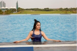 A person in a blue swimsuit at the edge of a pool with a clear view of a cityscape in the background.