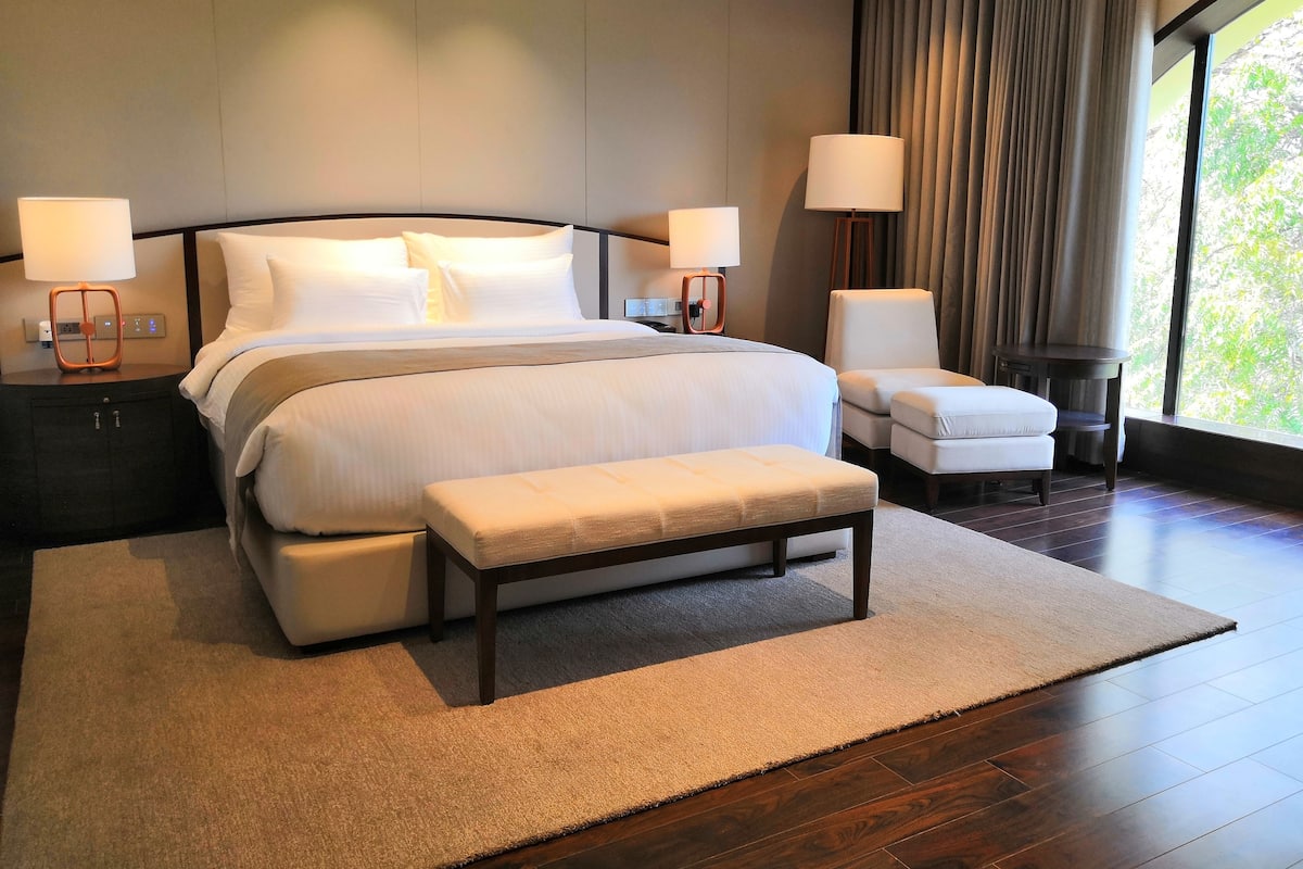 A modern hotel room with a large bed, two bedside tables with lamps, a bench, and a view of greenery through a large window.