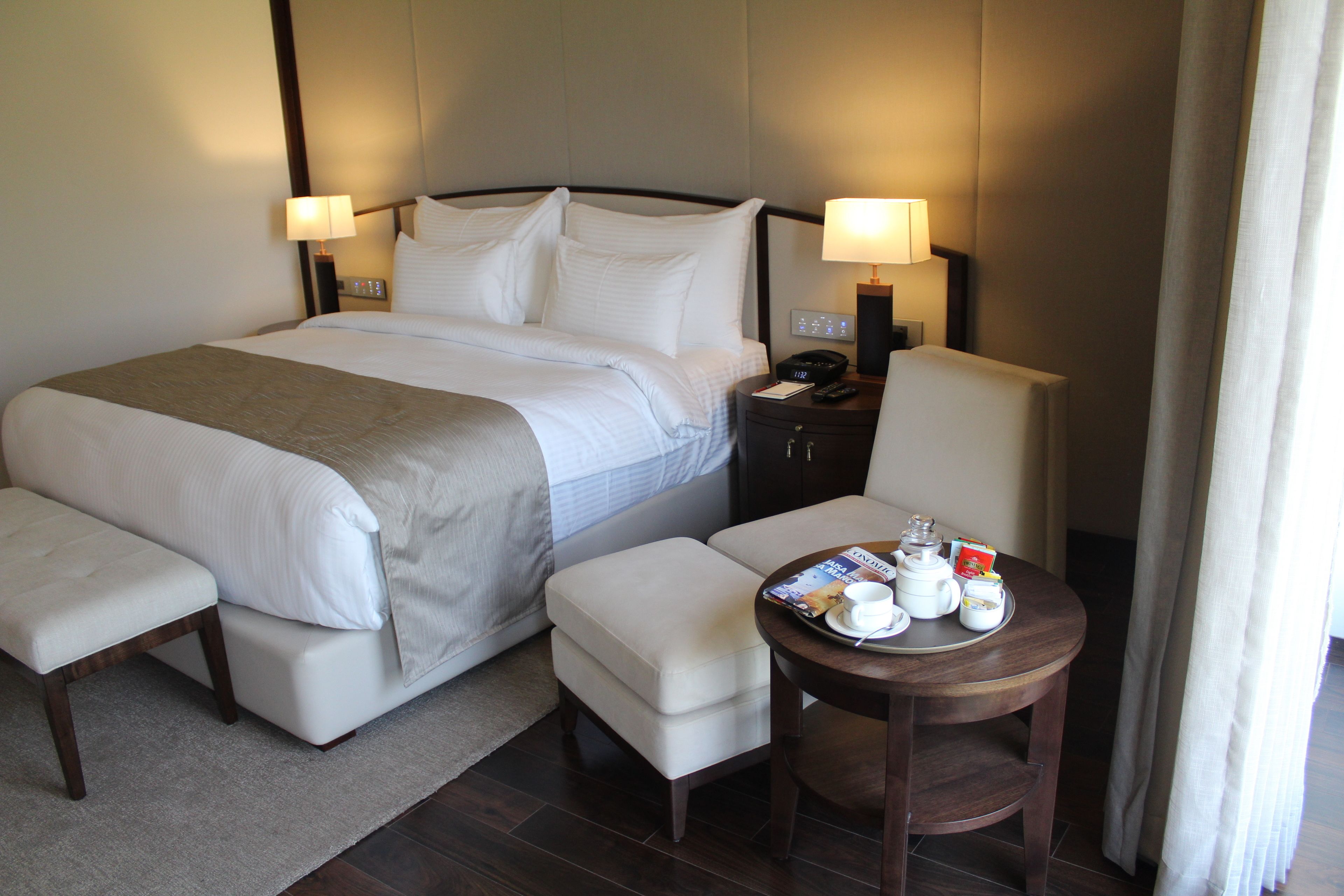 A hotel room with a large bed, bedside tables with lamps, a small round table with a tray of tea items, and a bench.