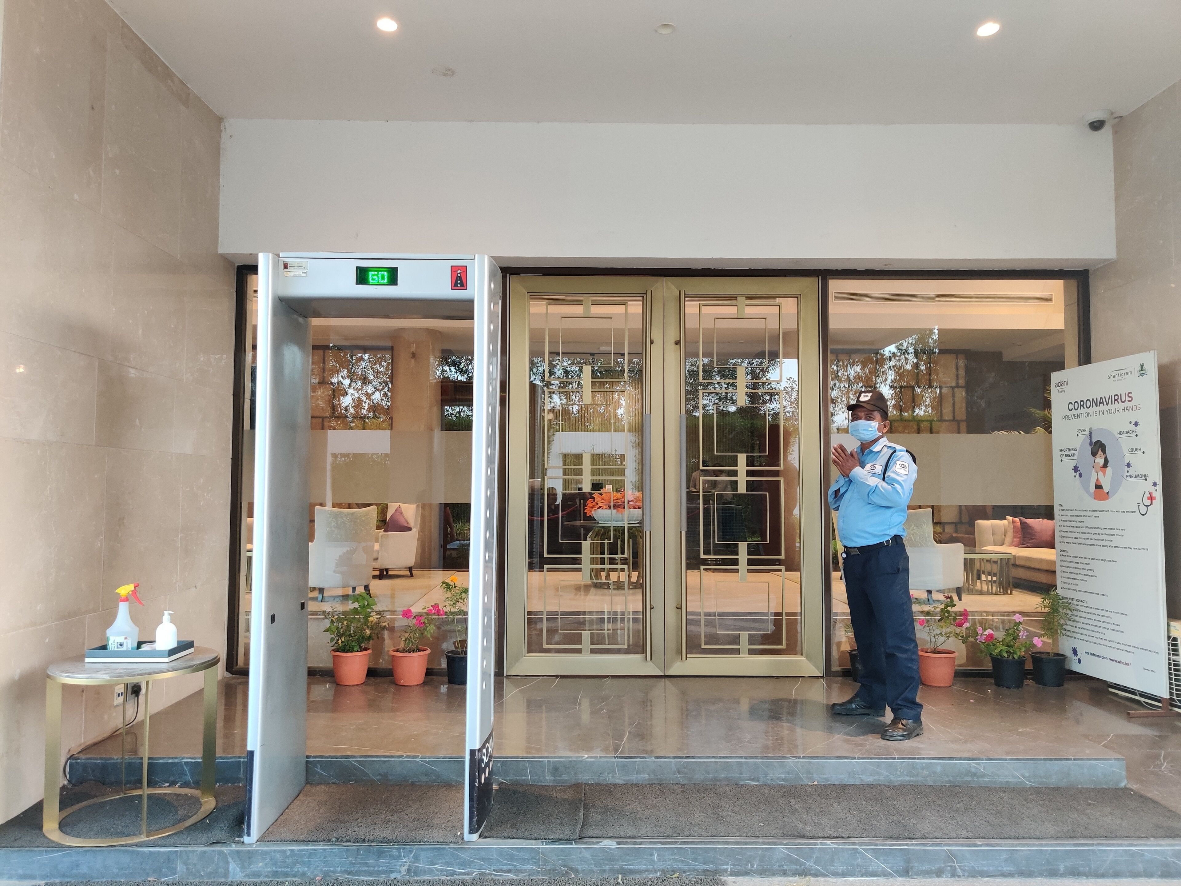 A hotel entrance with a digital display showing the temperature, a security guard, and a coronavirus prevention sign.