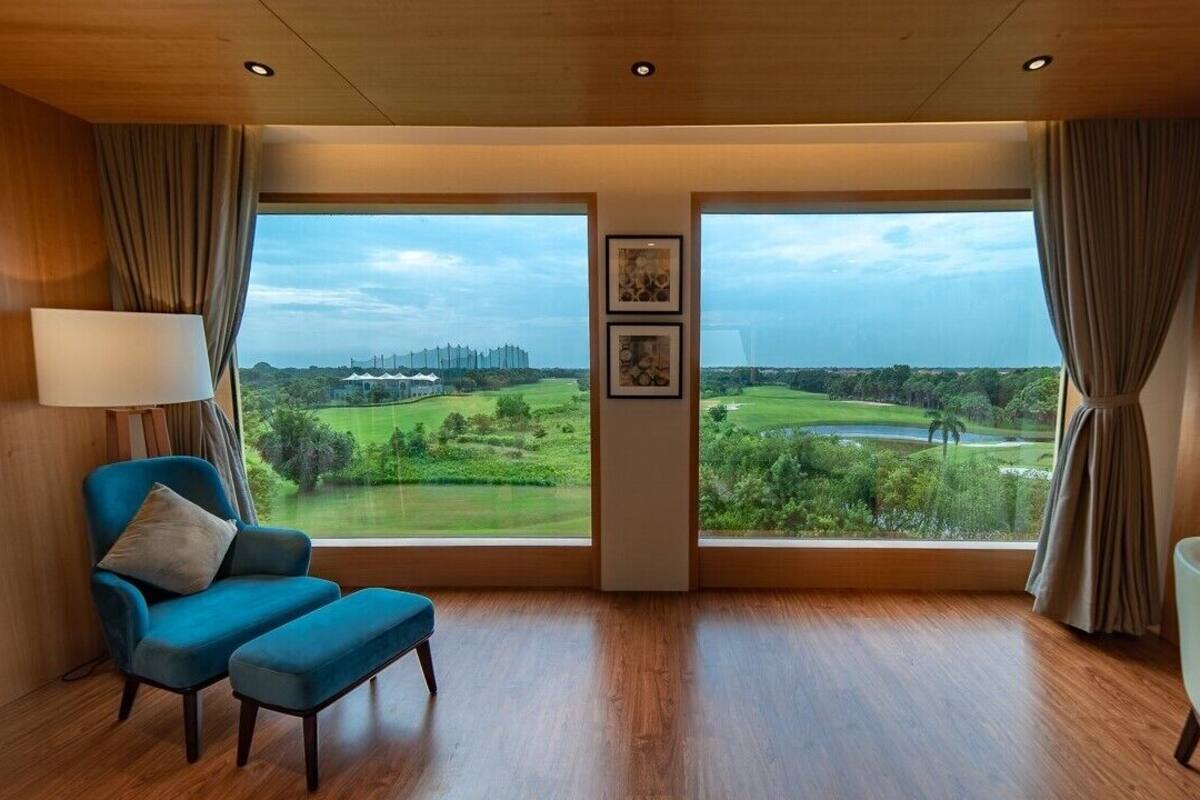A room with a large window offering a view of a golf course and distant buildings. There is a blue armchair and a small table with a lamp.
