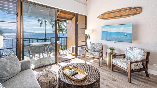 A spacious living area with a balcony, a round coffee table, and two patterned armchairs. - Maalaea Banyans 411 - Modern Renovation, Direct Ocean Front, Incredible View, AC (Wailuku)