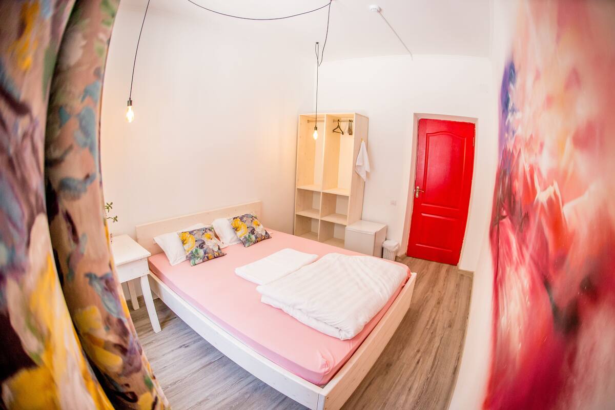 A bedroom with a pink bed, white pillows, a wooden wardrobe, a red door, and a white bedside table.