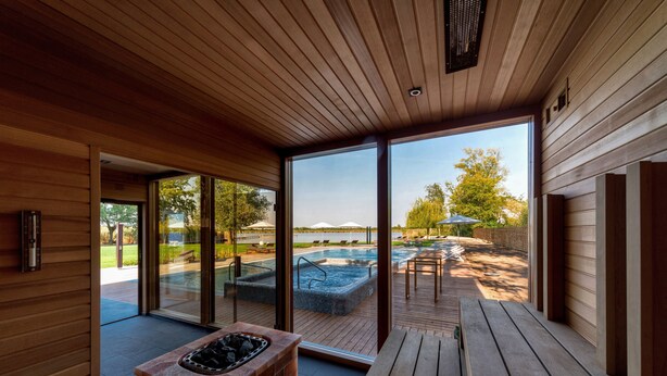 A modern indoor pool area with a wooden deck, a hot tub, and glass doors leading to an outdoor swimming pool. - Donautica Hotel & Restaurant (Fadd)