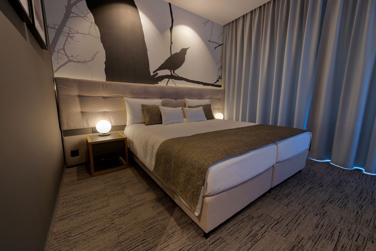 A modern hotel room with a large bed, a nightstand with a lamp, a wall mural of a tree and bird, and a window with curtains.
