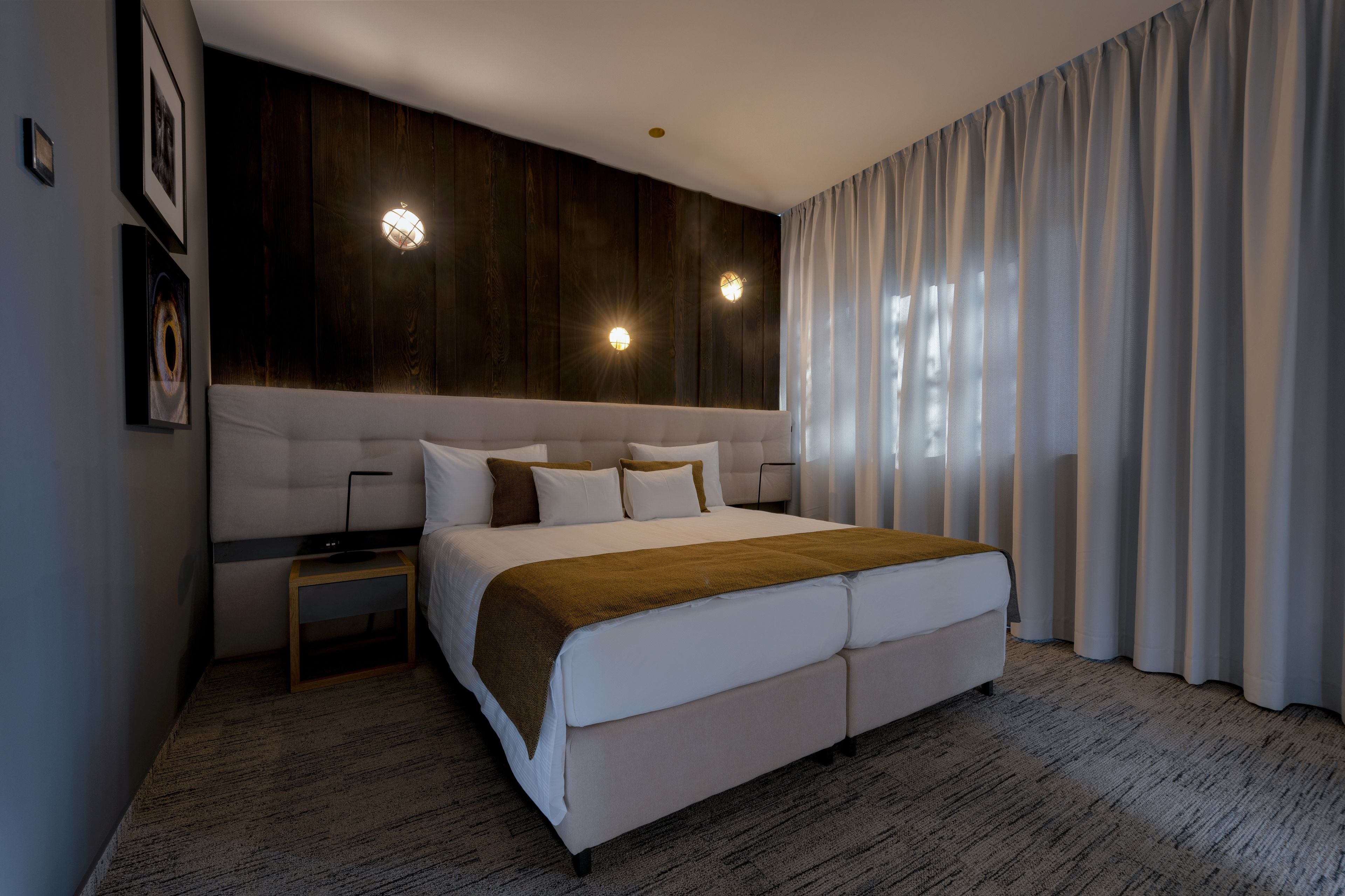A modern hotel room with a large bed, wooden panel wall, two bedside lamps, and a view of the outdoors through sheer curtains.