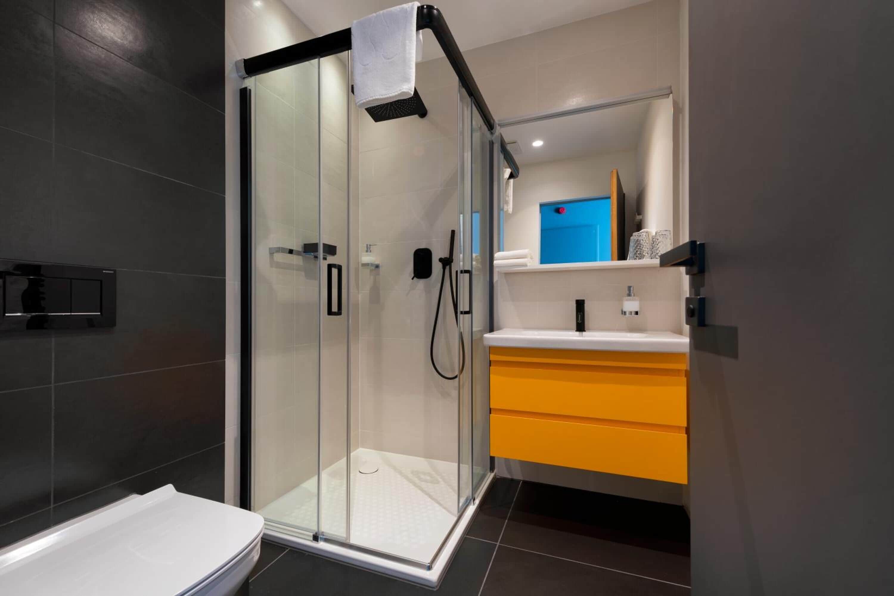 A modern bathroom with a glass shower enclosure, a white bathtub, and a yellow vanity with a blue mirror.