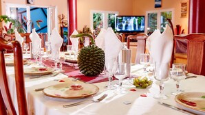A dining table set with white napkins, plates, and glassware, with a pineapple centerpiece and a television in the background. - Pinpin d'Amour (Saint-Philippe)