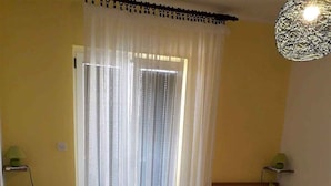 A room with two beds, a window with sheer curtains, a hanging light fixture, and a wooden floor.