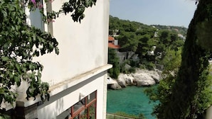 A white building with a balcony overlooking a turquoise body of water, surrounded by greenery and rocky terrain.