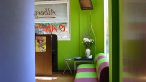 Una habitación con una pared verde, un sofá verde y púrpura a rayas, una mesita con un florero y una ventana con una persiana cerrada.