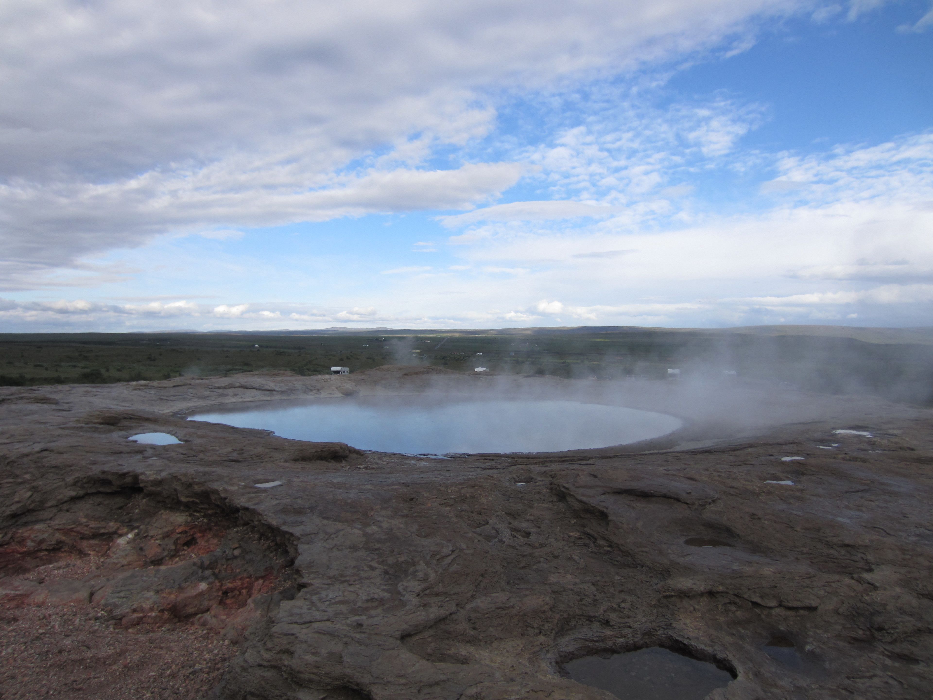 Une zone géothermique comprenant une source thermale circulaire, entourée d’un terrain rocheux et d’une végétation clairsemée.