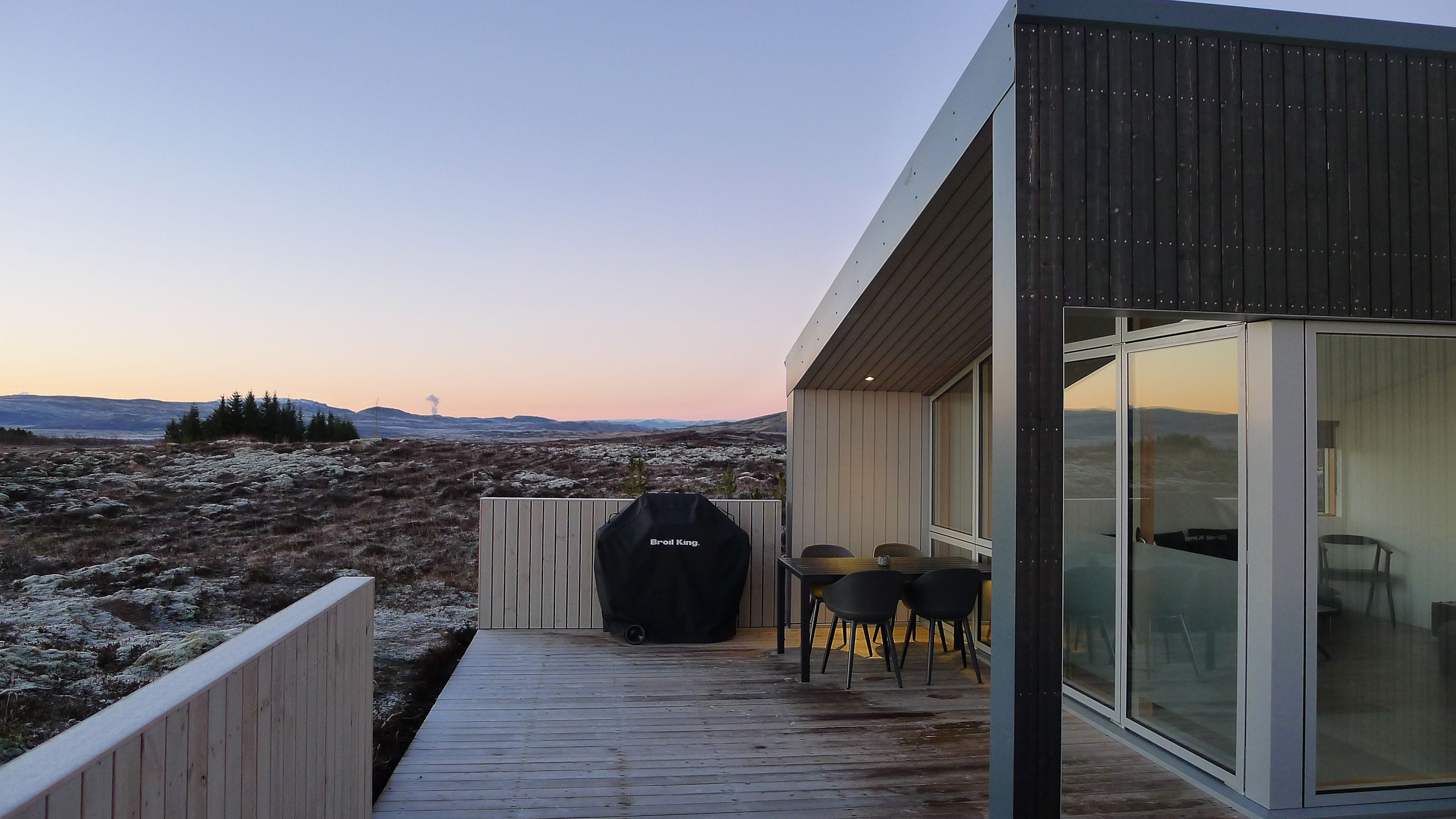 A modern outdoor deck with a grill, chairs, and a glass-enclosed room.