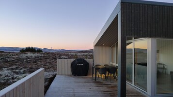 A modern outdoor deck with a grill, chairs, and a glass-enclosed room.