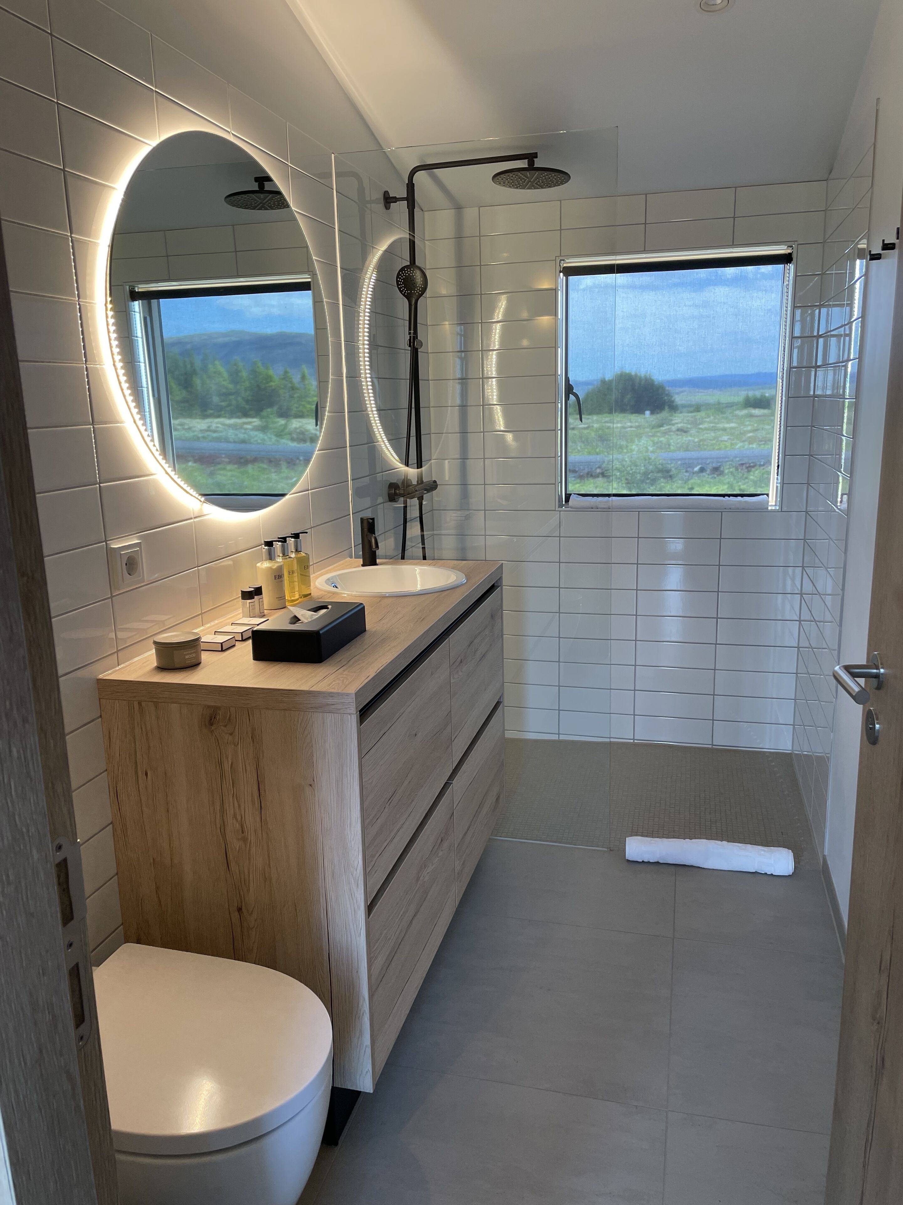 Un baño moderno con dos lavabos, un inodoro, una ducha y un amplio ventanal con vistas al exterior.