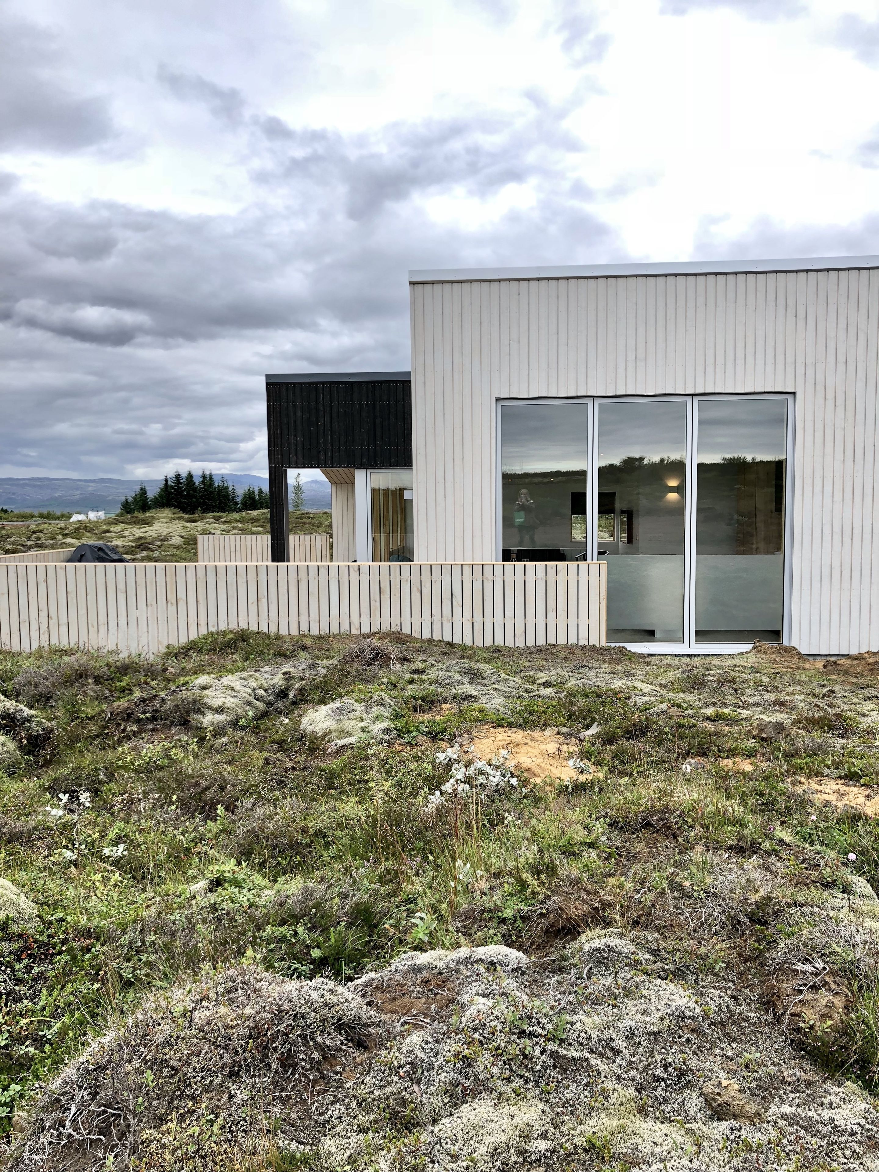 Een modern gebouw met grote glasramen en een houten omheining in een natuurlijke omgeving.