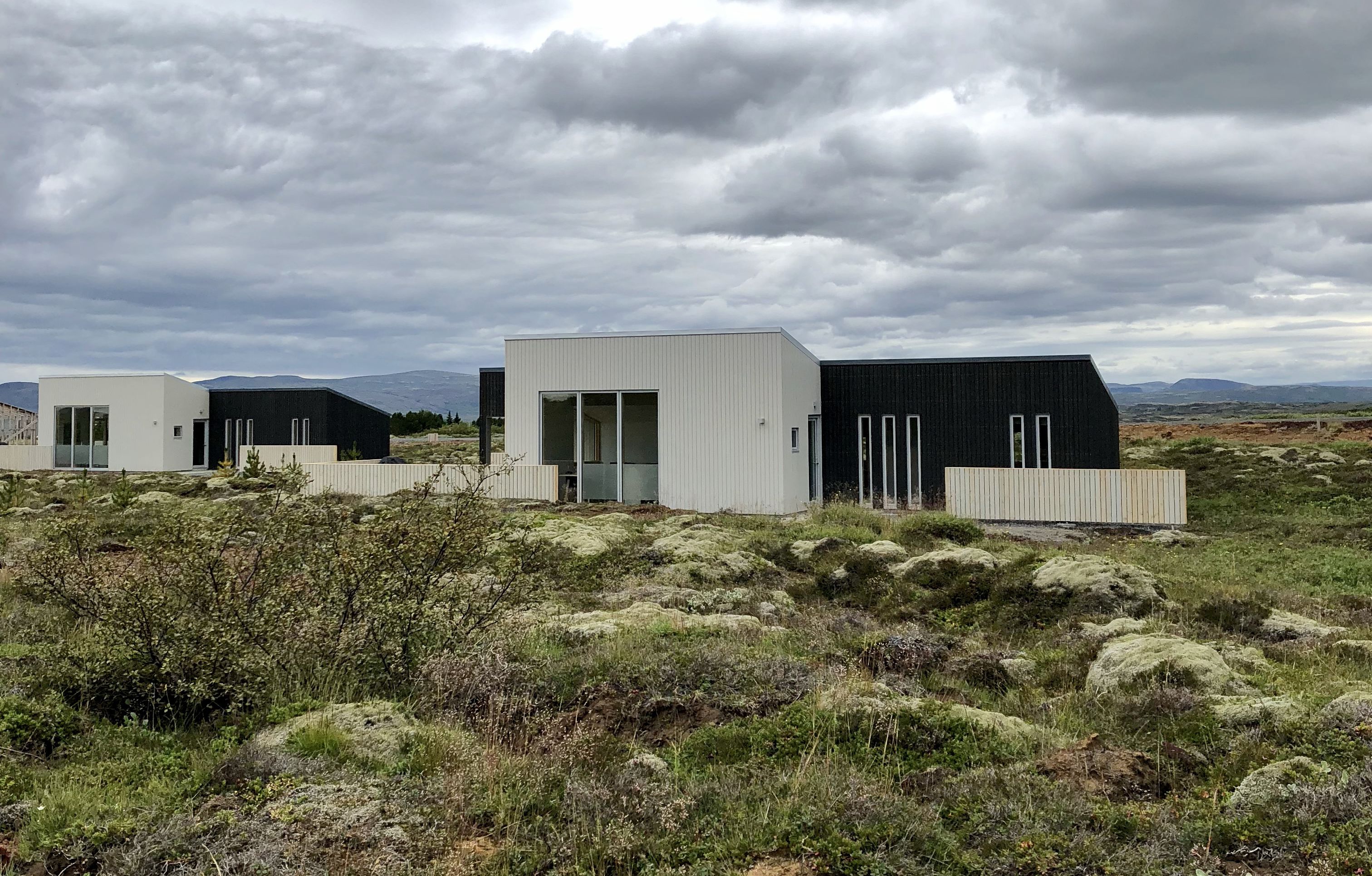 Un edificio moderno con techo plano y grandes ventanales, rodeado por un paisaje natural con vegetación escasa y montañas al lejano.