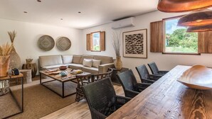 Een moderne woonkamer met een bank, salontafel en stoelen. Er hangen decoratieve kunstwerken aan de muur en is een groot raam met uitzicht op groen.