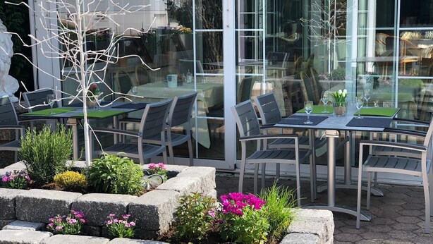 An outdoor seating area with tables and chairs, surrounded by stone planters with flowers. - Hotel Restaurant Angelo (Saarbrücken)