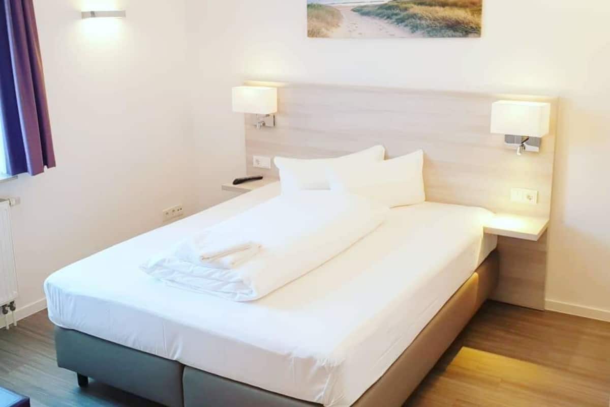 Una camera d'albergo con un letto, due lampade ai comodini, un quadro appeso al muro e una finestra con tende.
