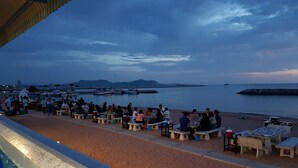 A beachside dining area with tables and chairs, a pool, and a view of the sea and boats.