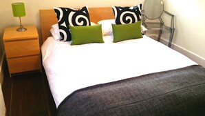 A neatly made bed with a white duvet and two green pillows, a wooden nightstand with a green lamp, and a round mirror on a clear glass table. - Victorian Terraced Apartments (London)