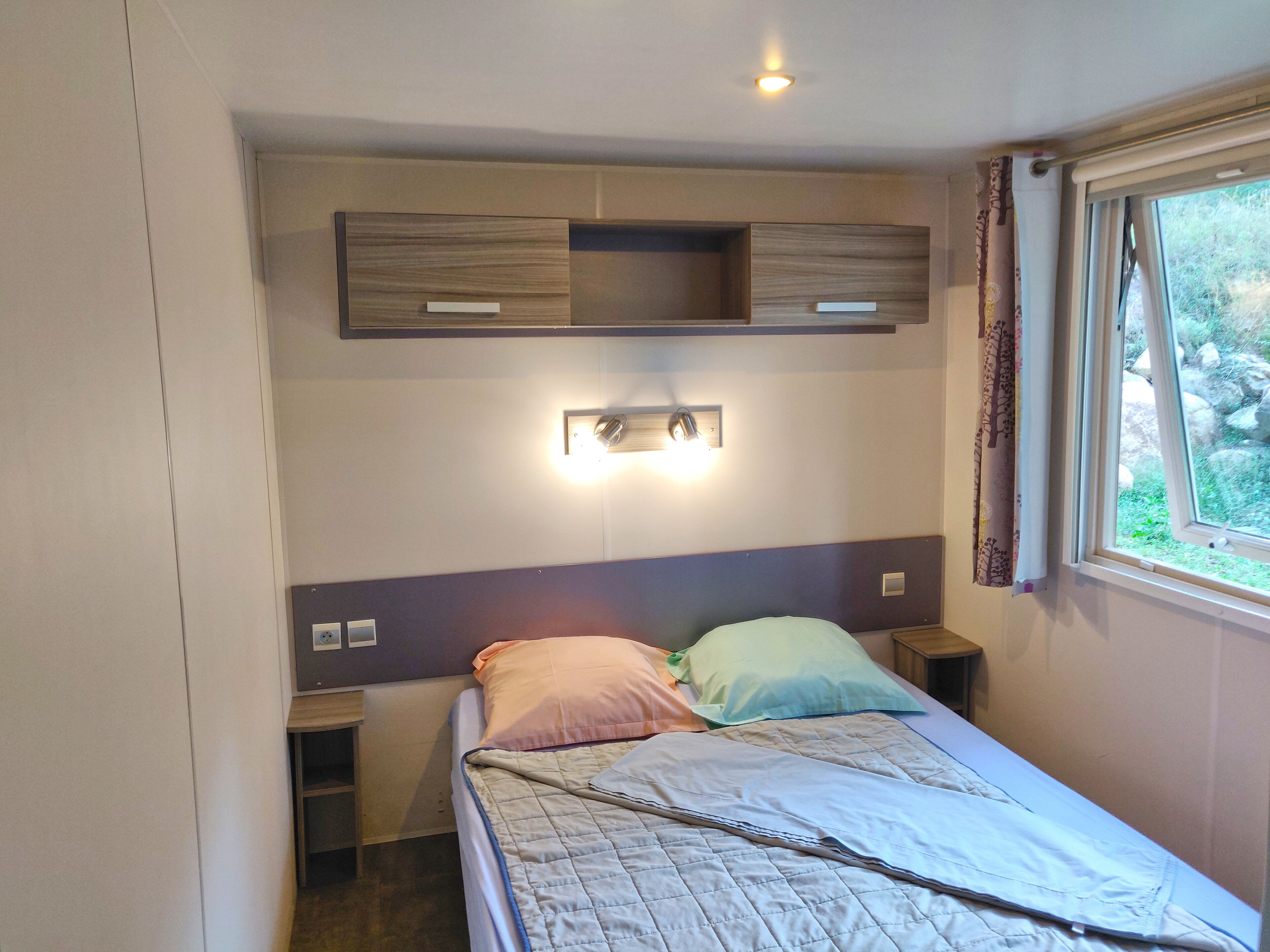 A single bed with a quilted bedspread, two pillows, a bedside table, a wall-mounted light, and a window with curtains.