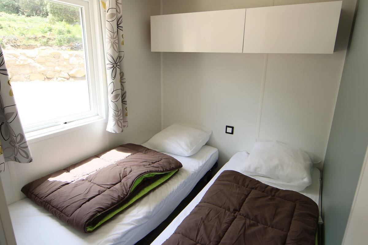A small, compact room with two beds, a window with curtains, and a white cabinet above the beds.