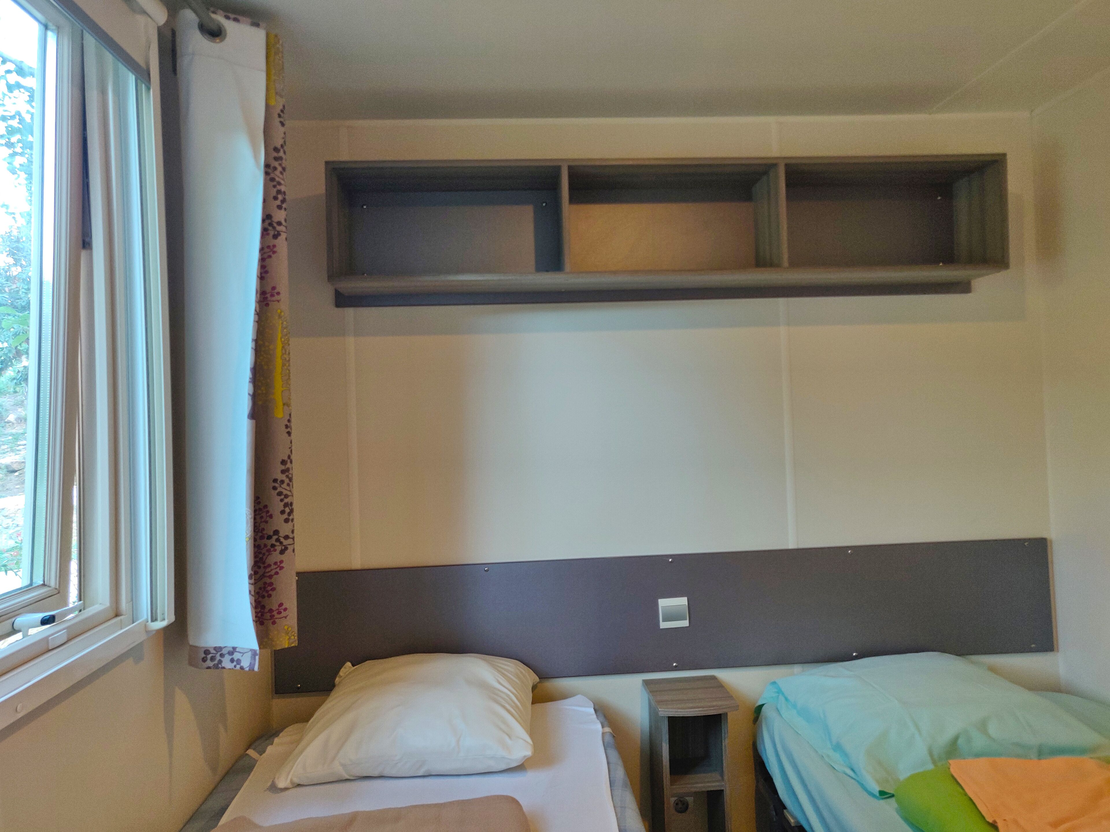 A small, single-bed room with a window, a bedside table, and a headboard with built-in shelves.