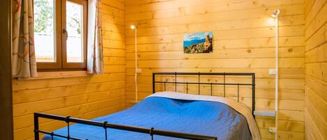 A wooden cabin room with a bed, a window with curtains, a wall-mounted light, and a poster.