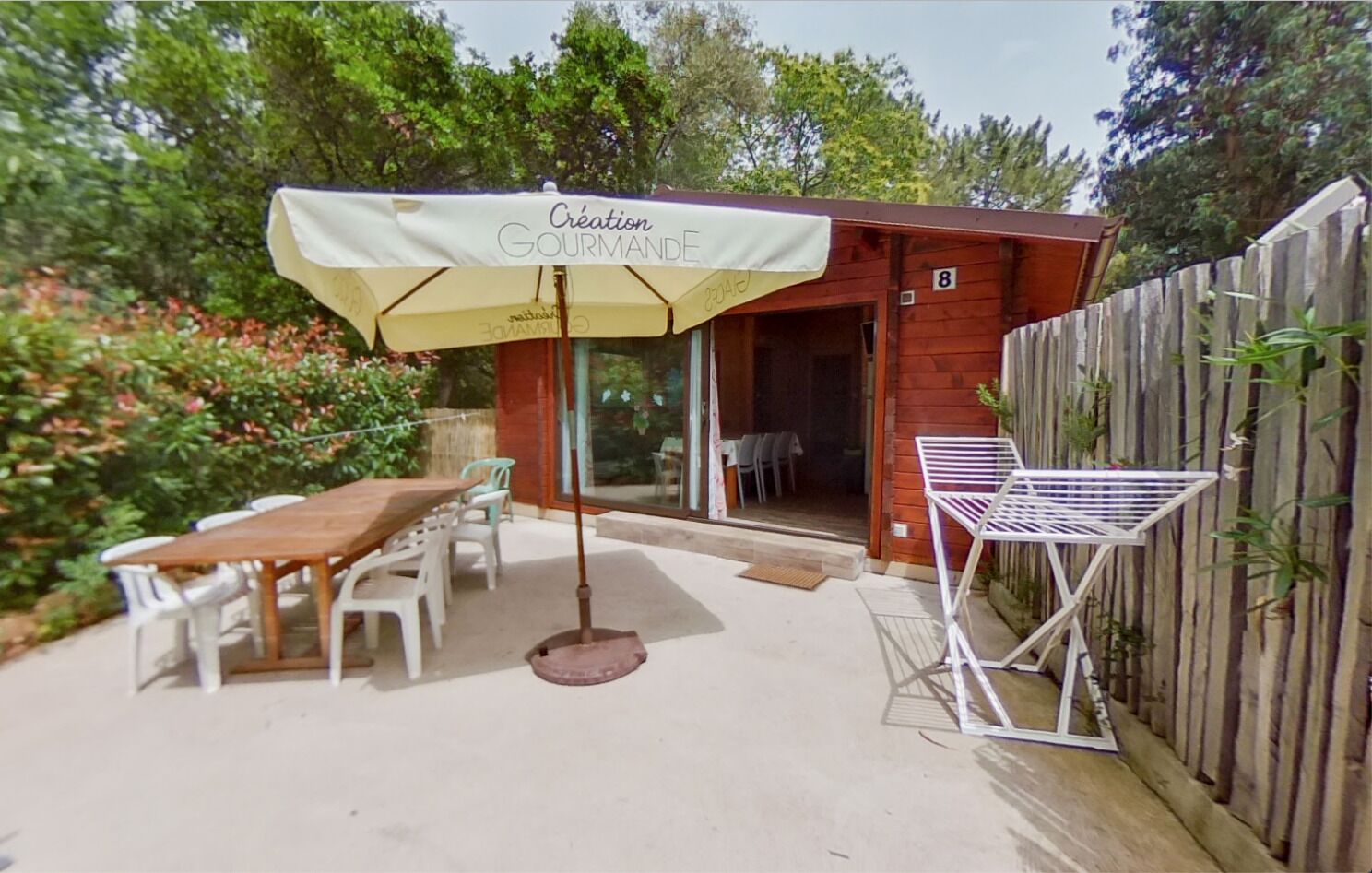 An outdoor patio with a wooden table and chairs, a covered umbrella, and a small wooden building with a glass door.