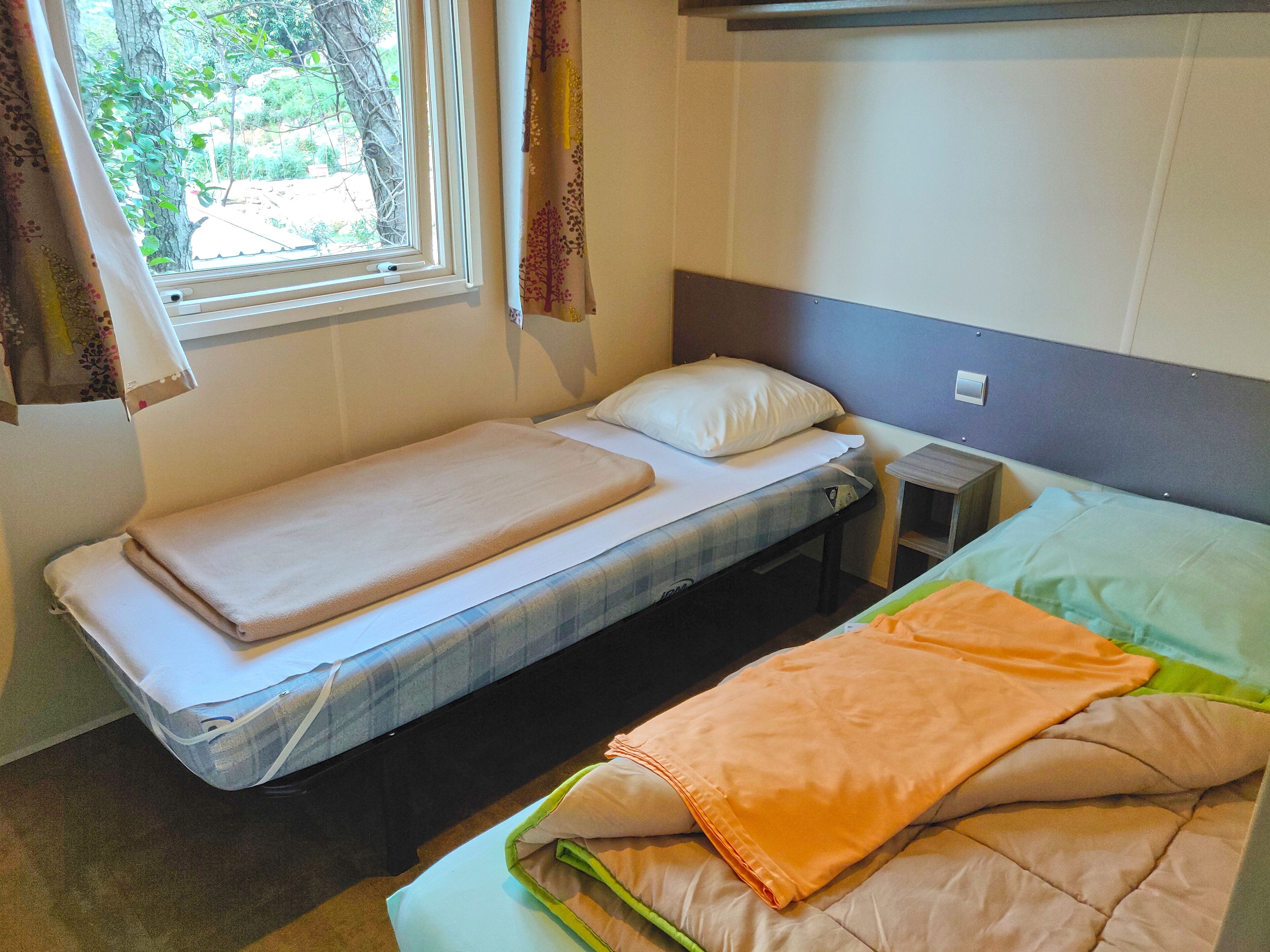 A single bed with a white mattress and a blanket, a bedside table, and a window with curtains.