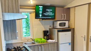 A compact kitchen with a gas stove, a microwave, and a refrigerator. A flat-screen TV is mounted above the stove, displaying an advertisement. - Funtana a l'ora (Ota)