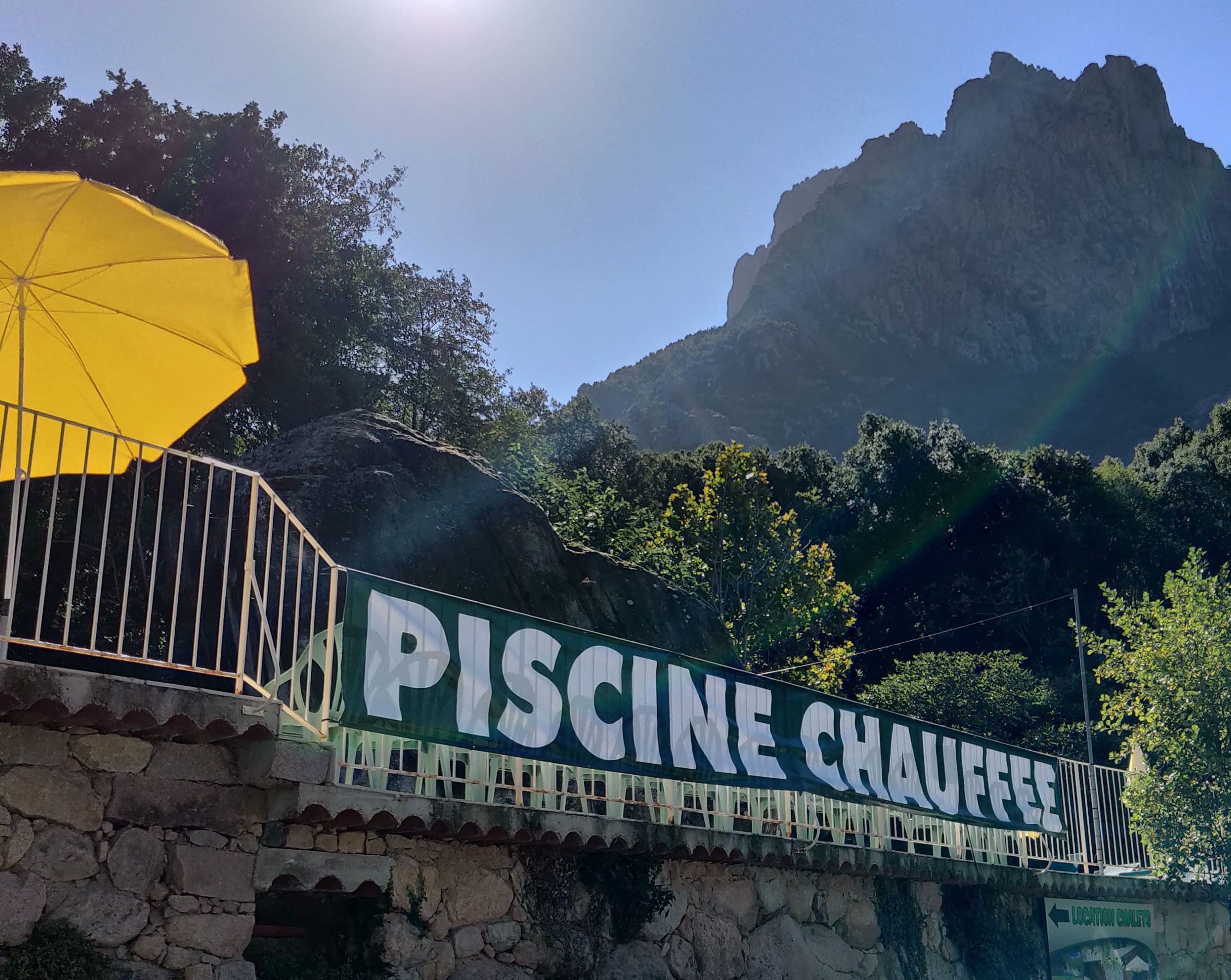 Señal de 'PISCINE CHAUEFFE' con un paraguas amarillo y vista a una montaña.