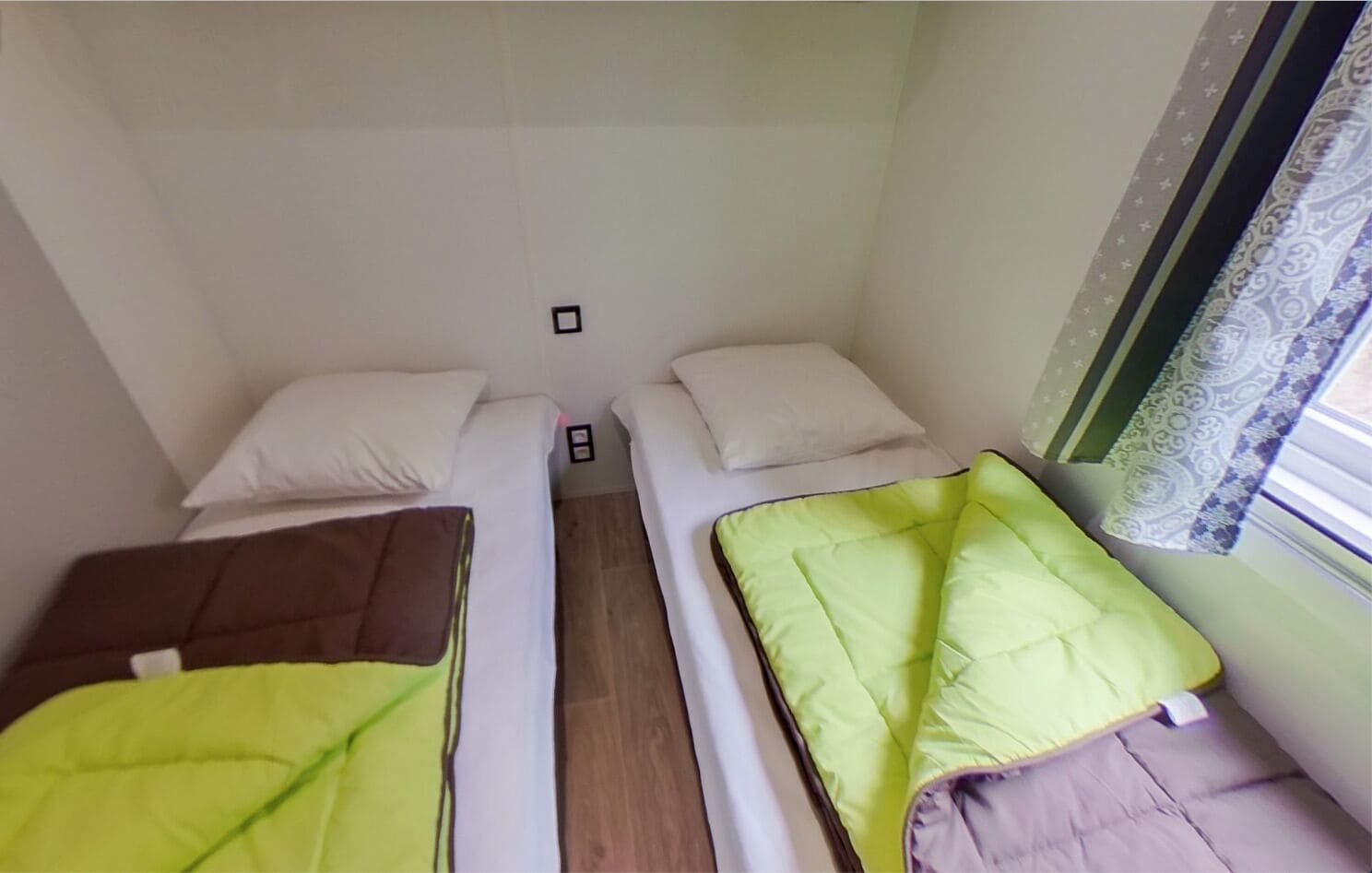A small, compact room with two single beds, each with a pillow and a blanket. There is a small table between the beds, and a window with patterned curtains.