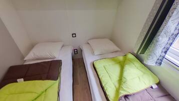 A small, compact room with two single beds, each with a pillow and a blanket. There is a small table between the beds, and a window with patterned curtains.