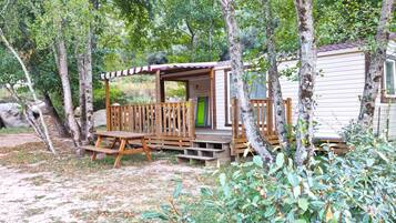 Een klein vakantiehuisje met een houten terras en een overdekte veranda, omringd door bomen en een stenen muur.