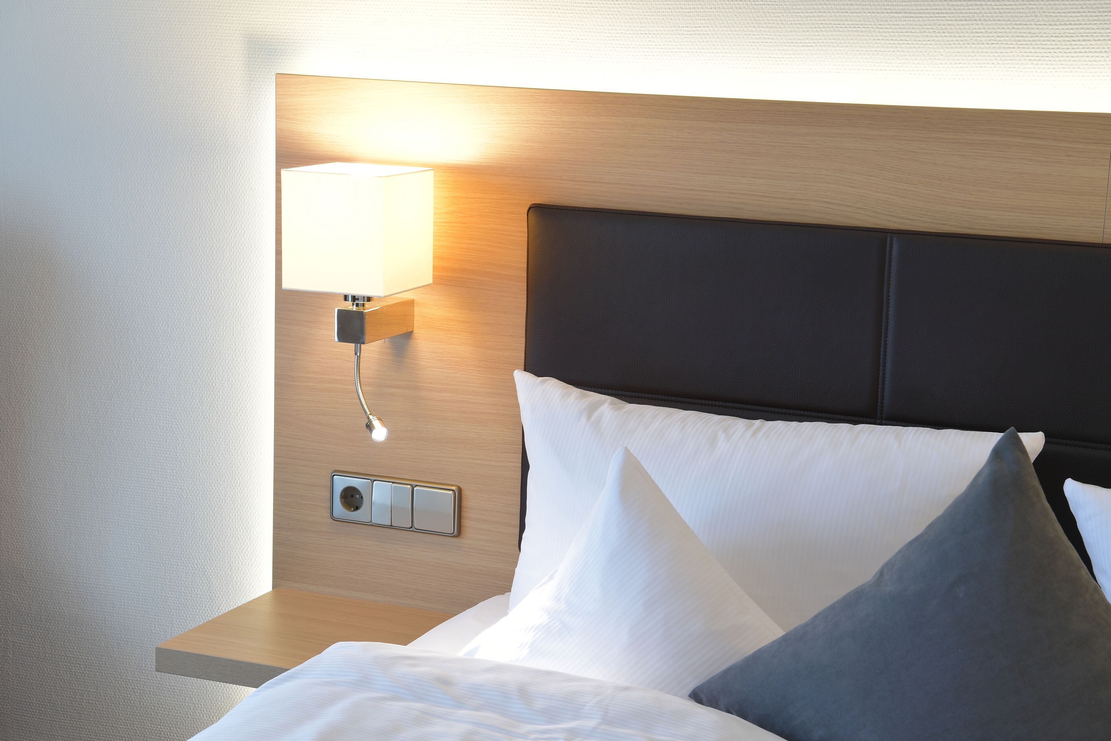 A neatly made bed with white and gray pillows, a wooden headboard, and a wall-mounted lamp.