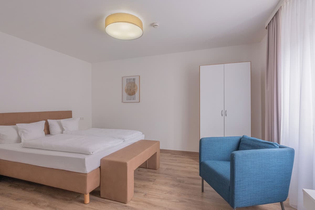 A hotel room with a bed, a bench, a blue chair, a wardrobe, and a ceiling light.