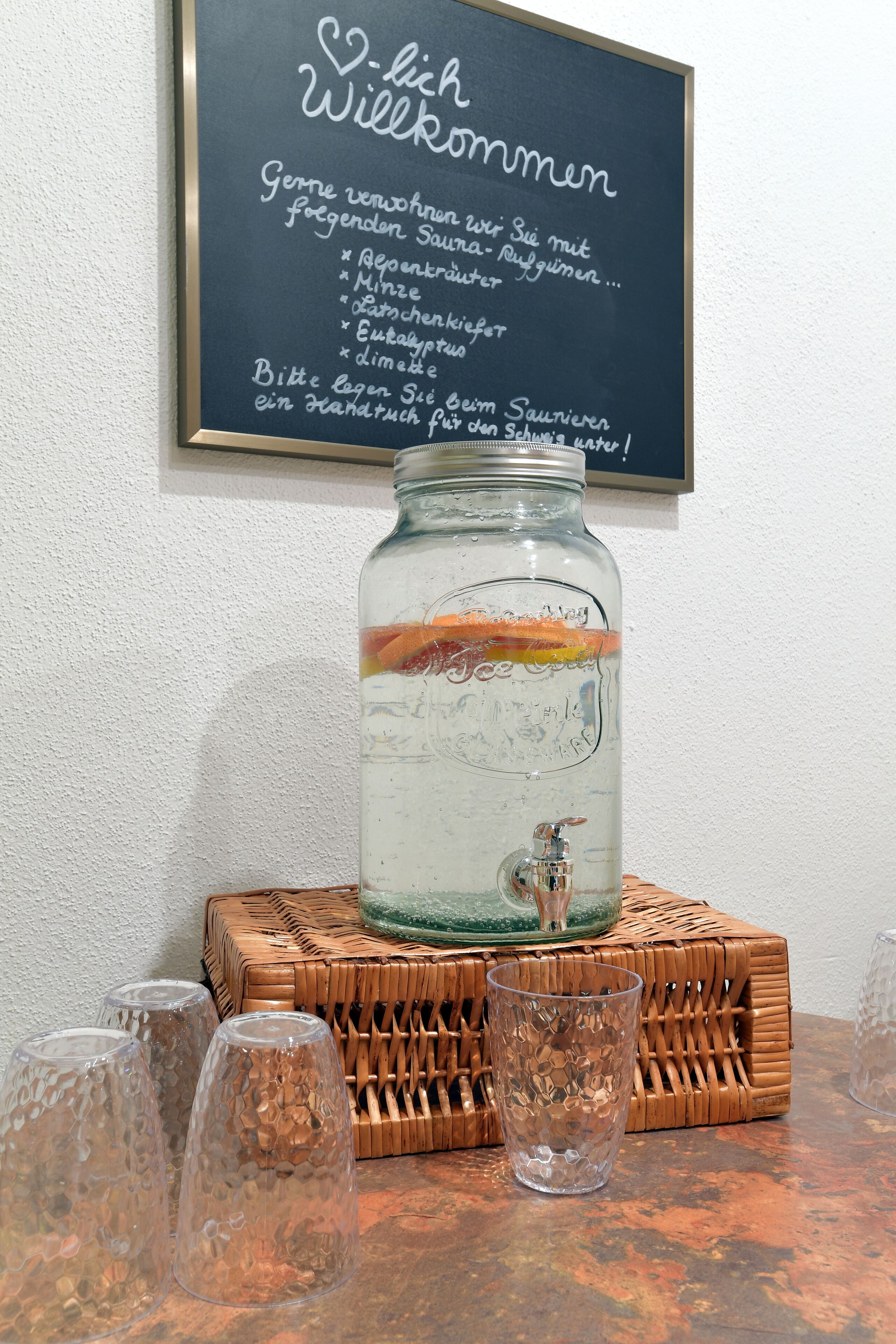 A glass jar with a water fountain, a chalkboard with a message, and three glasses on a table.