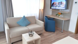 Una habitación de hotel moderna con un sofá color beige, una pequeña mesa de madera, una silla azul, un televisor montado en la pared y un escritorio con una lámpara y flores.