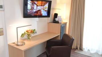 Ein Hotelzimmer mit Schreibtisch, Stuhl, Wandfernseher, Lampe und einem fensterbepflanzten Fenster.